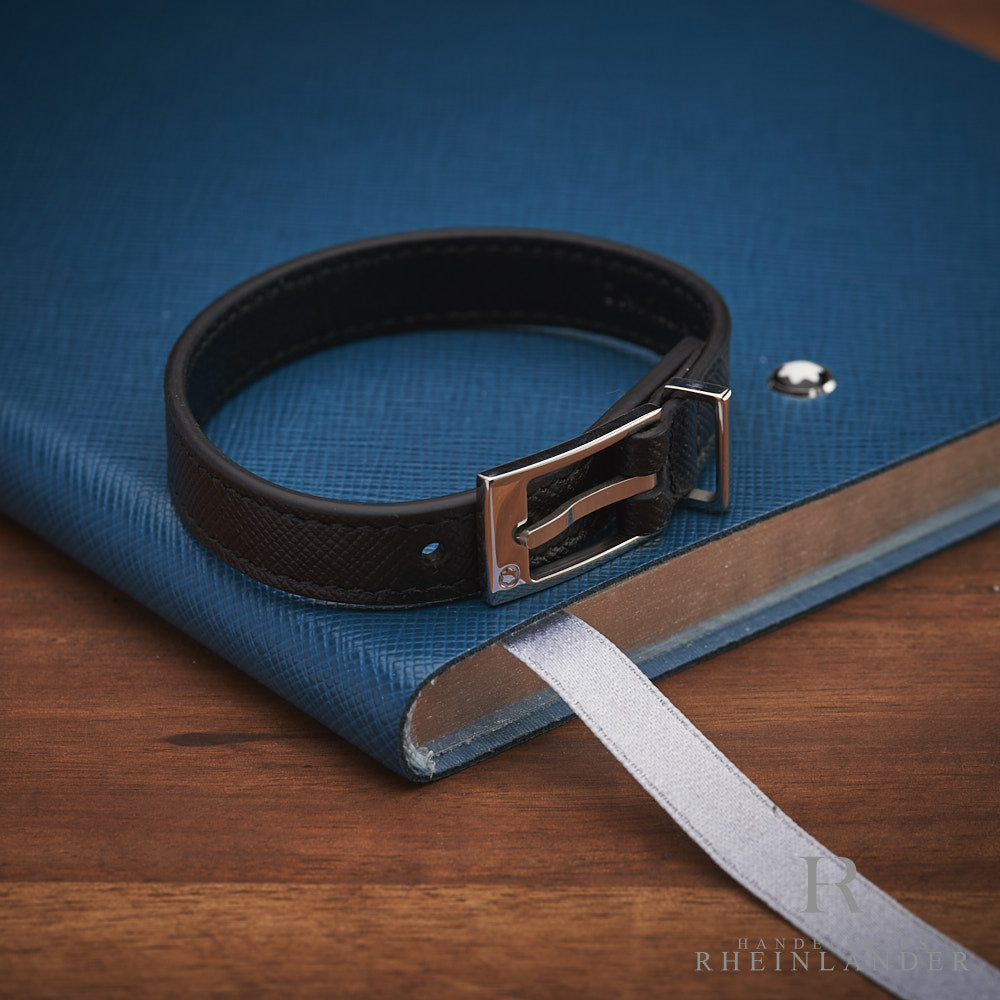 Schwarzes Lederarmband mit silberner Dornschließe auf blauem Notizbuch liegend, darunter Holztischplatte