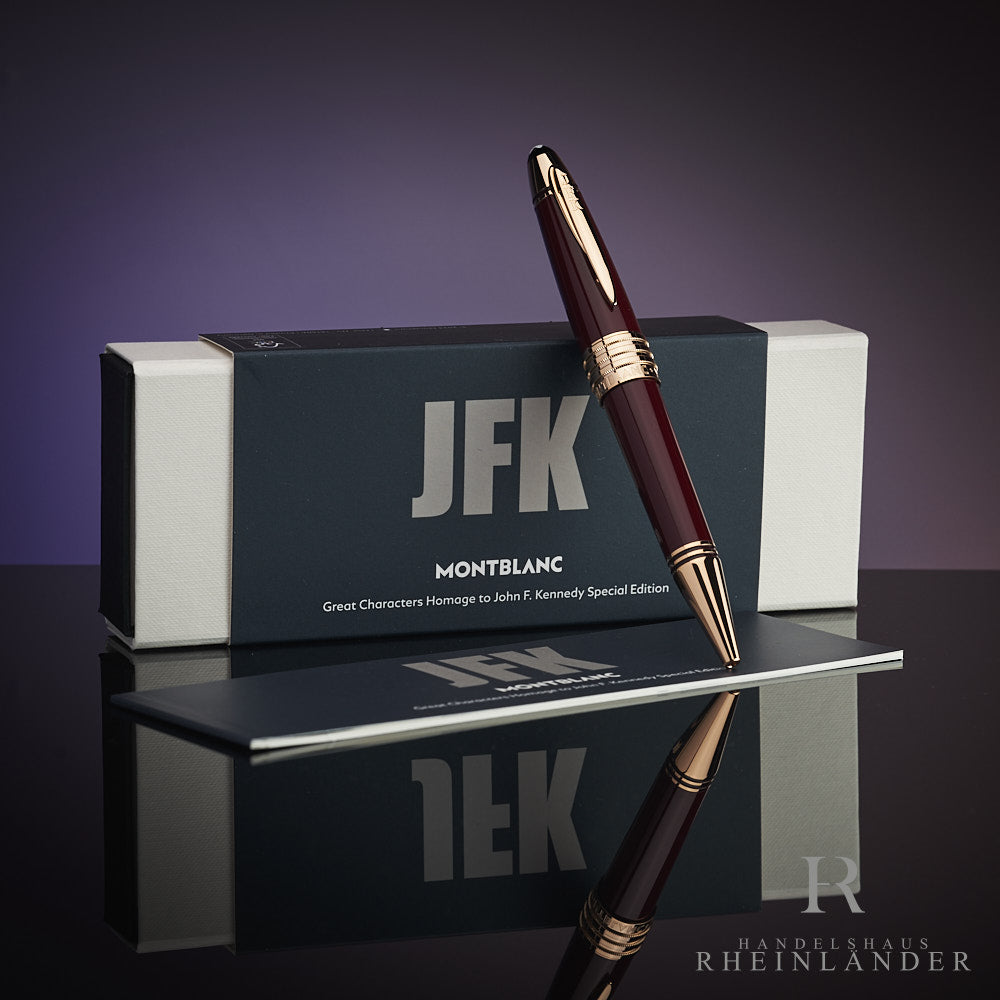 Dunkelroter Kugelschreiber lehnt an Originalbox mit JFK-Schriftzug. Unter dem Stift das Serviceheft, Hintergrund lila.