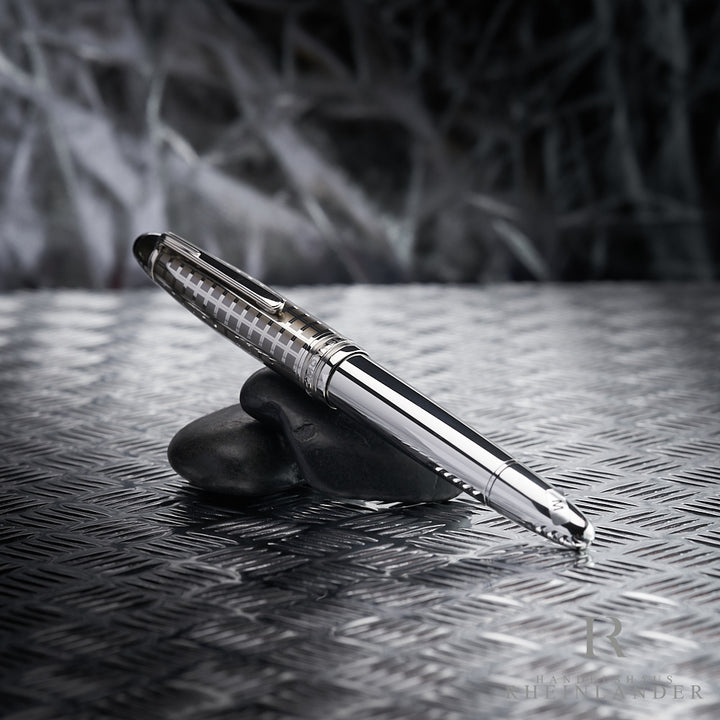 Silberner Füller mit gemusterter Kappe lehnt auf dunkelgrauen Steinen die auf Riffelblech liegen