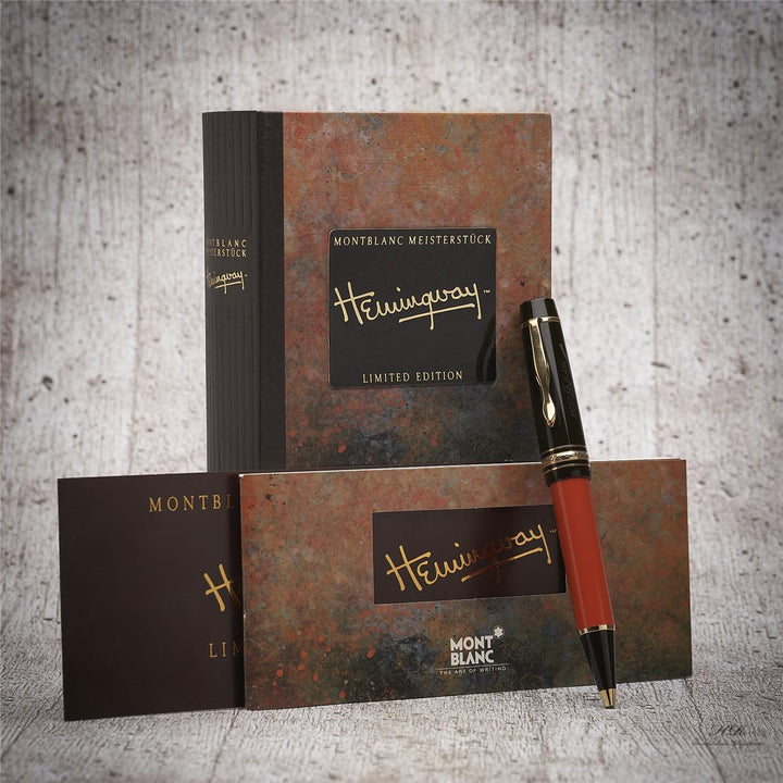 Roter Stift mit dunkler Kappe lehnt an Hemingway Buchbox und Serviceheft vor grauem Hintergrund in Holzoptik