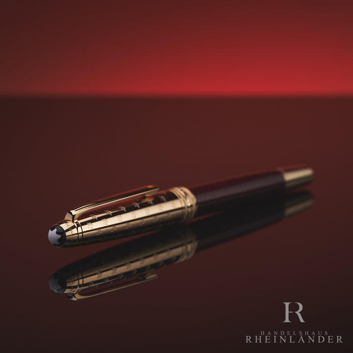 Burgundfarbener Rollerball mit goldener Kappe und Konus liegt mit Montblanc Logo zur Kamera auf spiegelnder Oberfläche vor rotem Hintergrund 