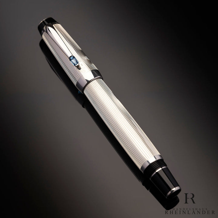 Geschlossener, silberner Rollerball mit blauem Edelstein auf dem Clip liegt auf anthrazitfarbenem Hintergrund