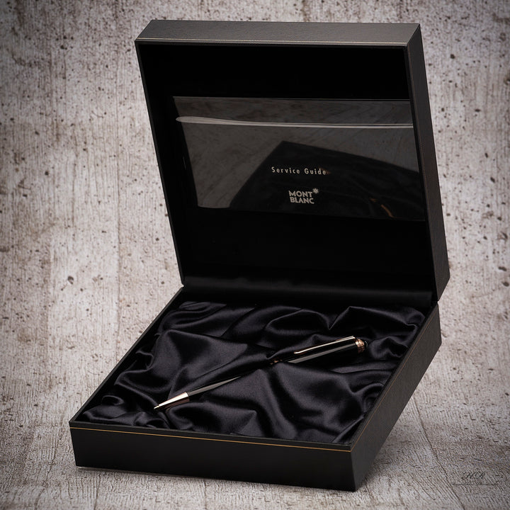 Schwarzer Drehbleibstift von Montblanc mit roségoldenen Beschlägen in offener Originalbox liegend vor grauem Holzoptik-Hintergrund fotografiert
