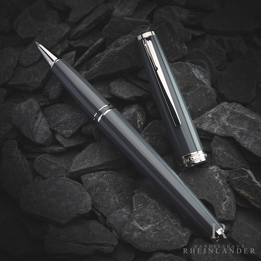 Grauer PIX Rollerball von Montblanc, daneben graue Kappe mit silbernen Beschlägen, auf dunkelgrauem Steinboden fotografiert